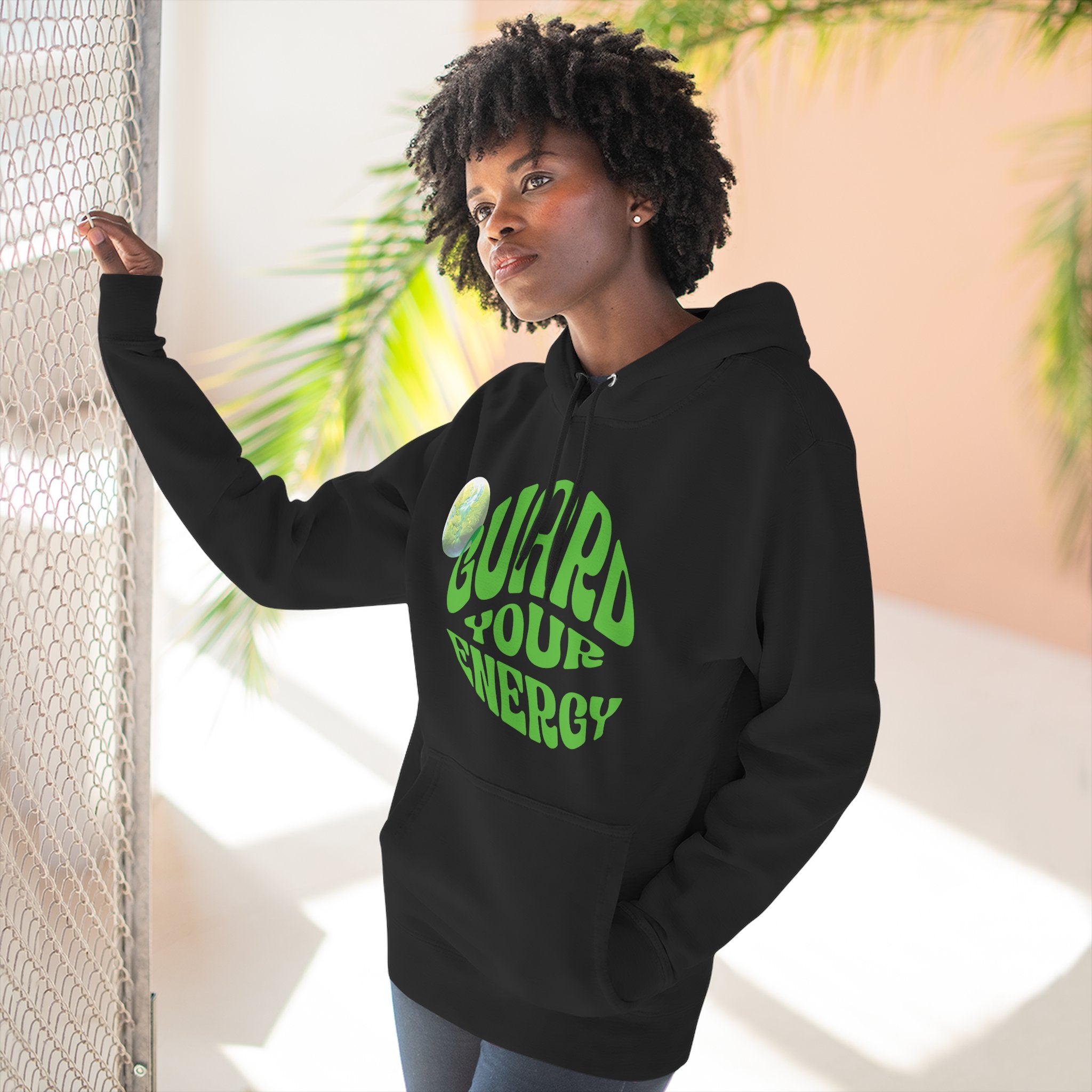 woman wearing black Guard Your Energy Hoodie with green graphic text and kangaroo pocket standing by a window with sunlight and plants in the background