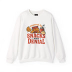 White crewneck sweatshirt with "powered by SNACKS AND DENIAL" graphic featuring chips, cookie, drink, and dumpling